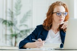 Serious experienced female coach makes records in notepad, focused at screen of laptop computer, has