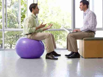 Businessman on exercise ball in meeting with colleague using laptop computer in exercise studio, sid